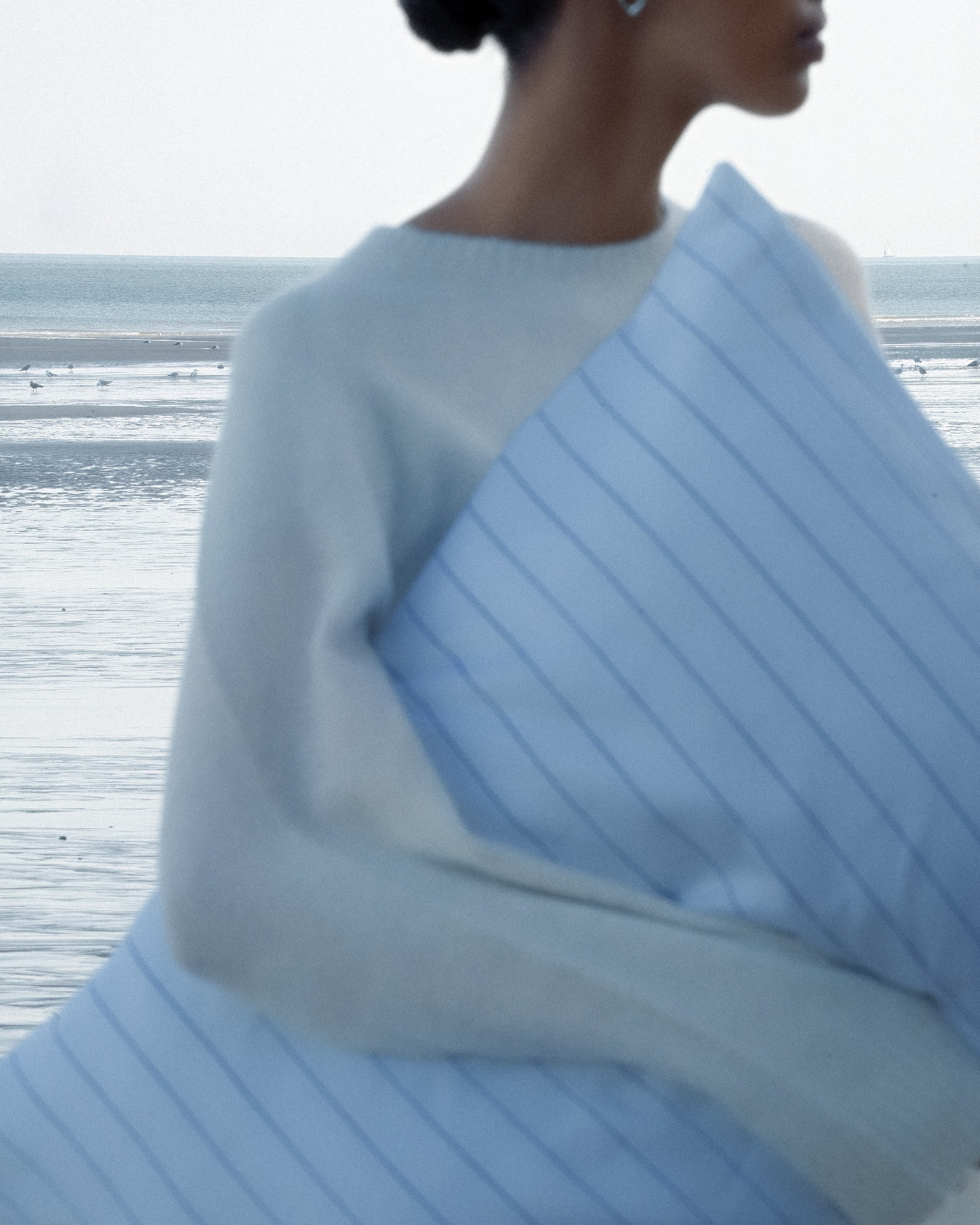Blurry image of a woman carrying a soft pinstripe pillow, standing in front of the blue ocean.